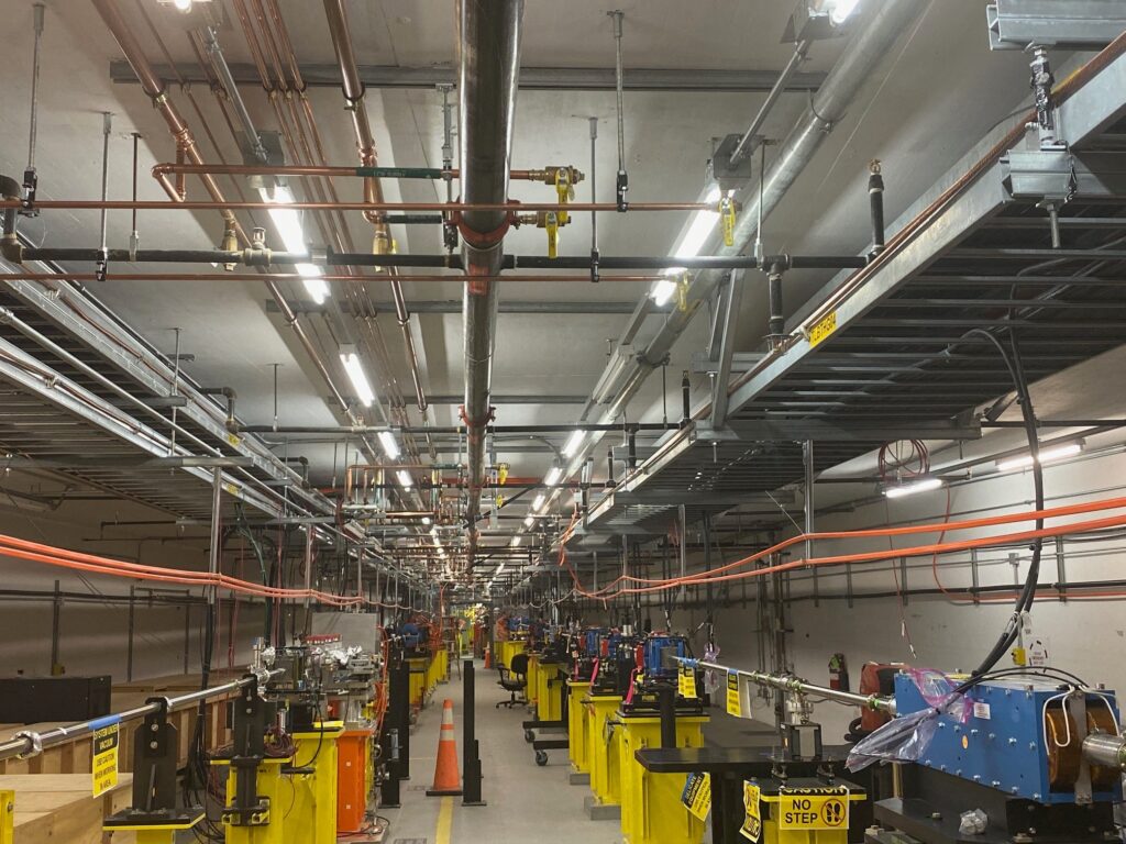 A large industrial workshop with rows of colorful machines on both sides. Overhead, there are numerous pipes and cables. The floor is marked with safety warnings, and a single orange traffic cone is visible in the aisle.