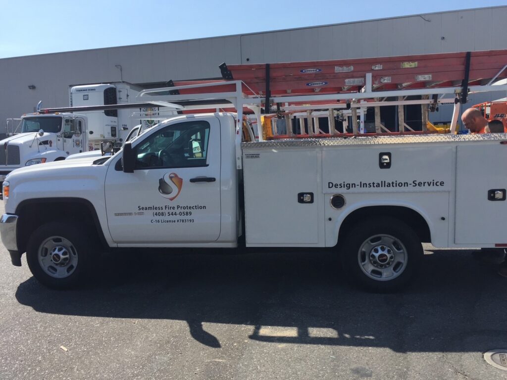 A white utility truck with Seamless Fire Protection branding is parked in a lot. The truck carries red ladders on top and has markings indicating Design-Installation-Service. Other vehicles are seen in the background.