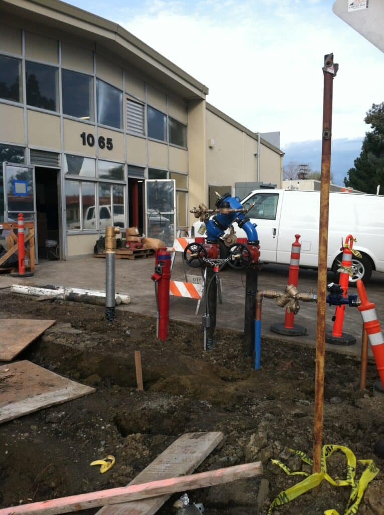 Construction site in front of a commercial building. Pipes and a blue valve structure are exposed in a dug area. Traffic cones and caution tape surround the site. A white van is parked nearby, and the building has large windows and an open door.