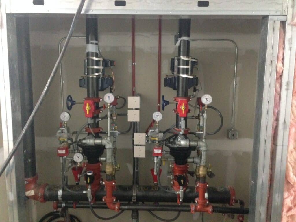 A complex plumbing setup featuring two sets of industrial pipes and valves mounted on a wall. Each set includes gauges and red and black components. The pipes are secured within a metal frame.