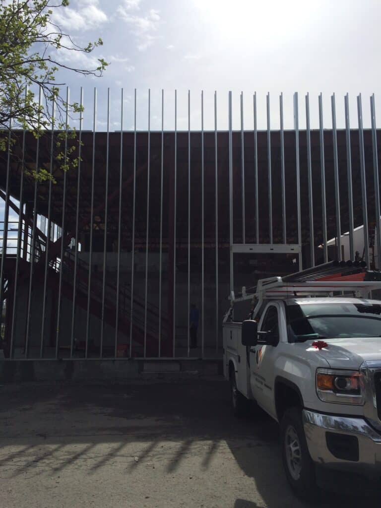 A construction site with vertical metal beams in front of a partially built structure. A white utility truck with a ladder on top is parked in the foreground. The sky is partly cloudy, and there are some tree branches on the left.