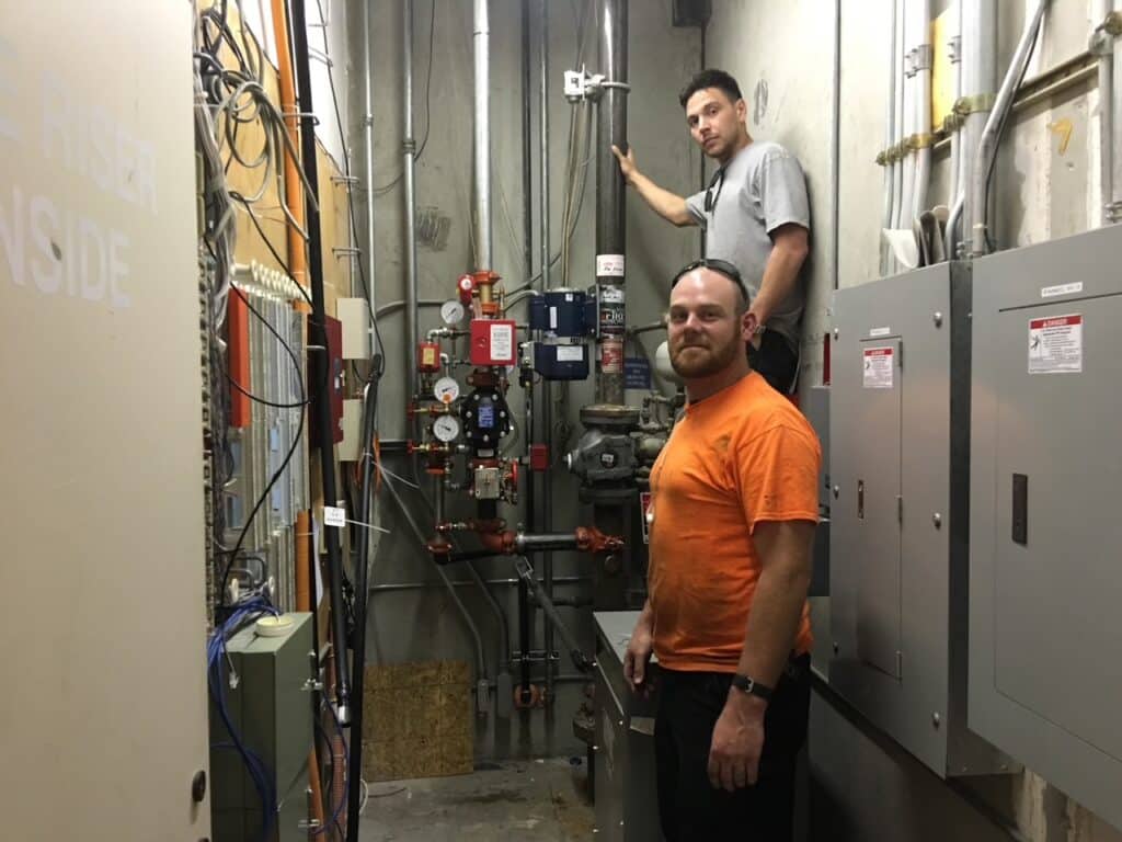 Two men are in a utility room surrounded by electrical panels and pipes. One is standing on a step ladder working with tools on the wall, while the other stands on the ground, facing the camera.