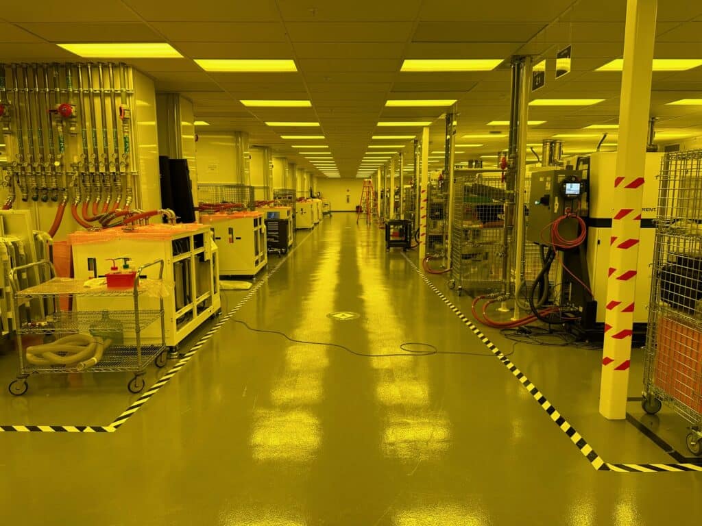 A bright, yellow-lit laboratory or cleanroom with polished floors and various industrial equipment along the walls. The room is organized with safety tape marking pathways and caution signs visible. Ceiling lights illuminate the spacious environment.