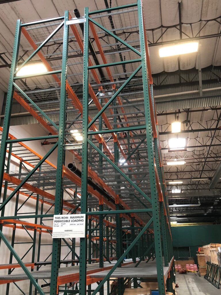 Warehouse interior with tall metal storage racks. The structure has green vertical supports and orange horizontal beams. Overhead lights illuminate the space. A sign on the rack mentions maximum permissible loading. Cardboard and wooden pallets are on the floor.