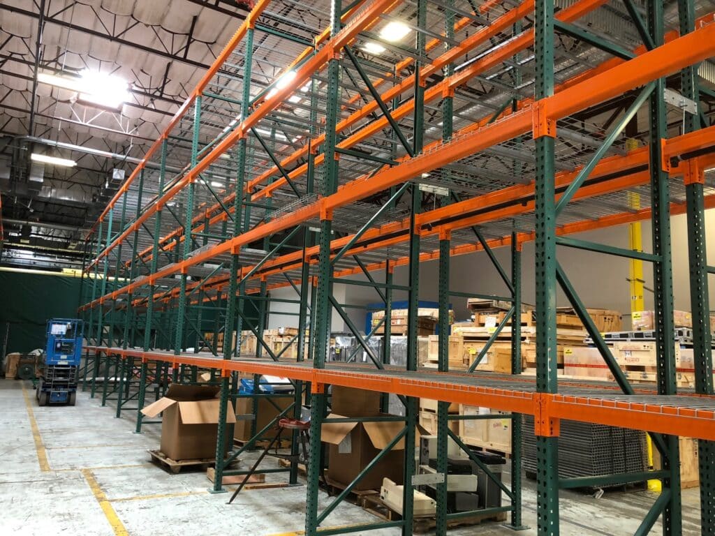 Warehouse interior with empty orange and green metal shelving units. Cardboard boxes and wooden crates are scattered on the floor. Overhead lighting illuminates the industrial space. A blue mechanical lift is visible in the background.