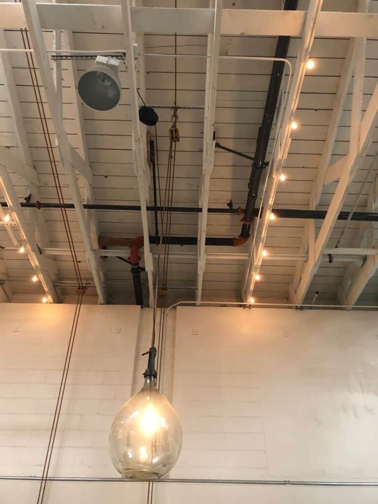 A large, spherical light bulb hanging from a ceiling with exposed beams and pipes. Additional smaller lights along the beams create a rustic, industrial look. The walls are painted white, complementing the minimalist, modern aesthetic.