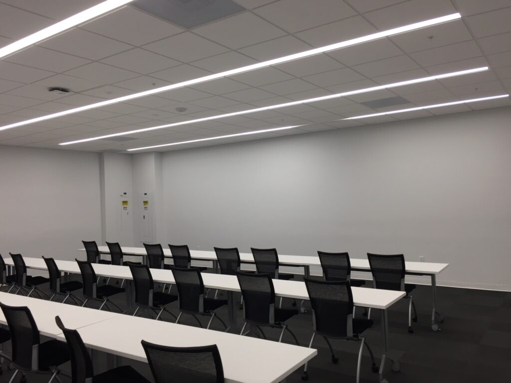 A modern classroom with white walls and ceiling, featuring rows of white desks and black office chairs. The room is well-lit with rectangular ceiling lights and has a minimalist design. Theres a blank wall at the front of the room.