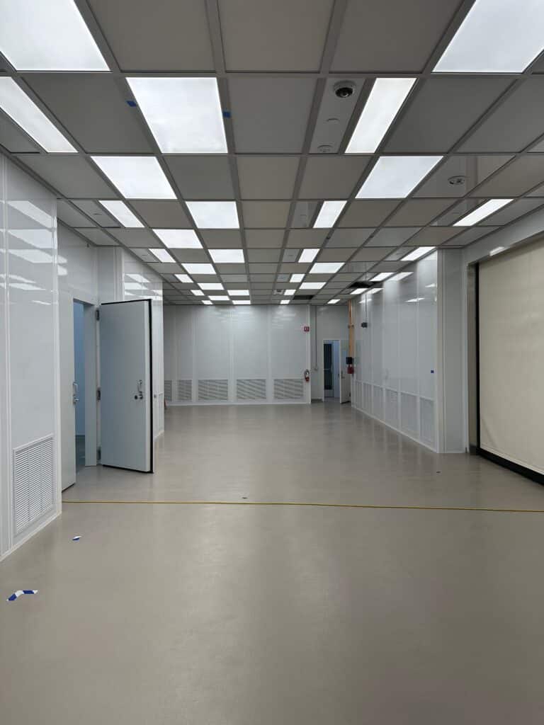 Empty room with a beige floor and white walls, illuminated by several rectangular fluorescent lights on the ceiling. There is an open blue door on the left and a closed white door on the right. The space appears to be clean and spacious.