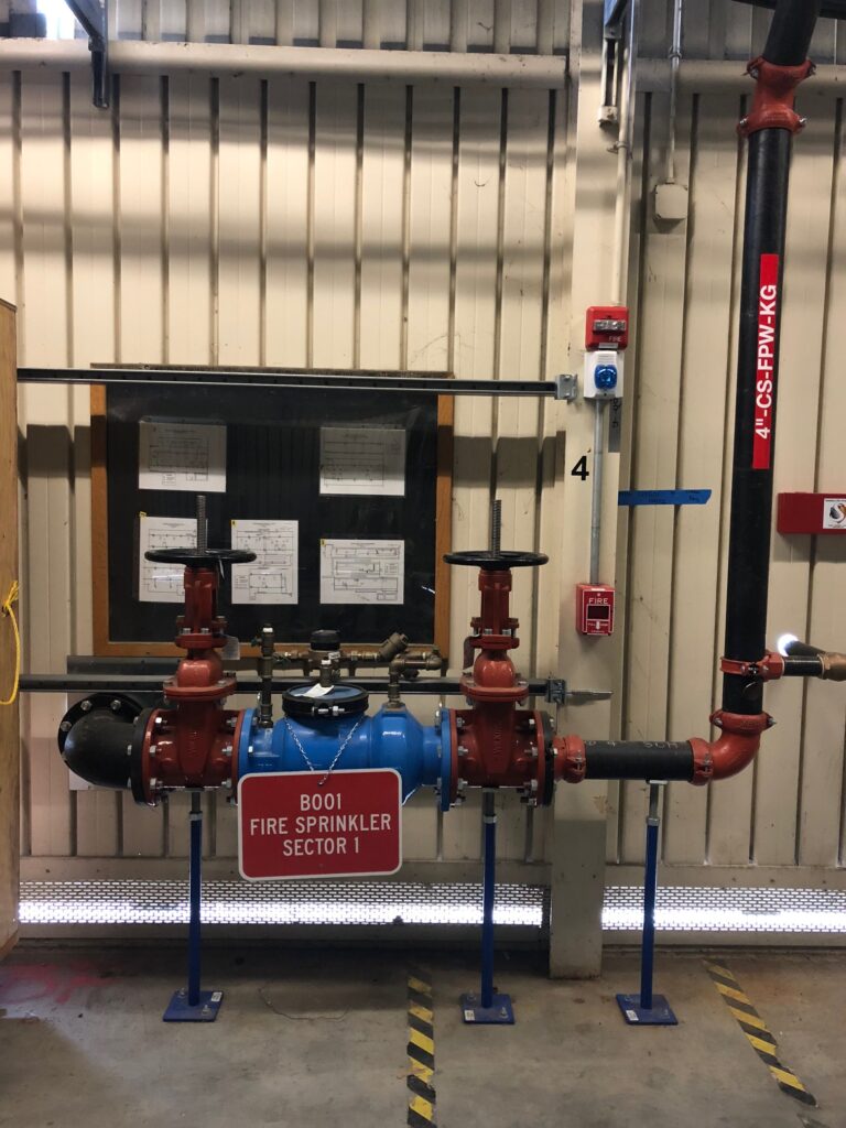 Fire sprinkler system labeled B001 Fire Sprinkler Sector 1 inside an industrial building. It features red and blue pipes and valves with a background of plans on a bulletin board and a striped safety floor marking.