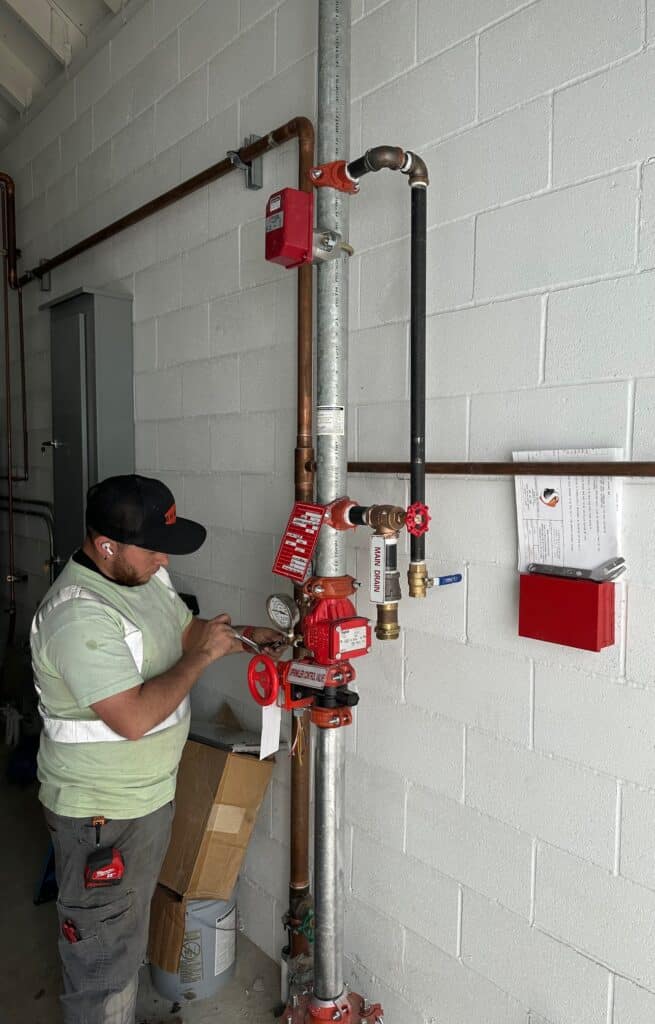 A man in a cap and safety vest is inspecting a fire sprinkler system with gauges and red valves, attached to a wall. He is holding a wrench and notes. A clipboard with papers is mounted on the wall nearby.