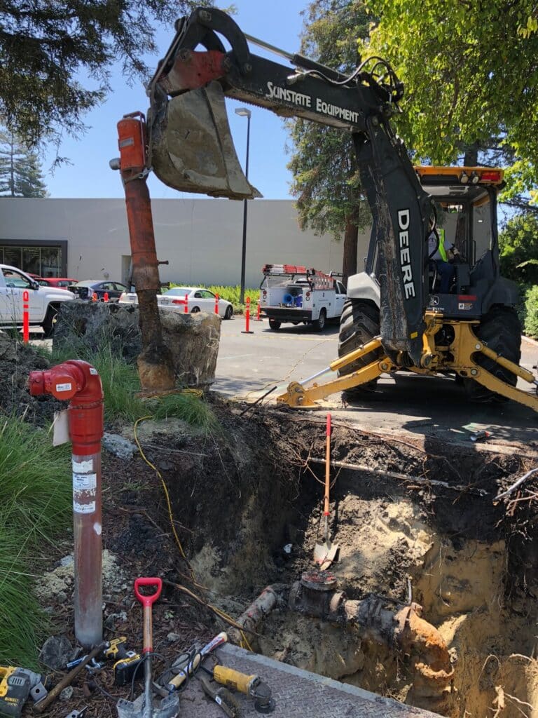 A construction site with a backhoe from Sunstate Equipment digging near a sidewalk. The area is marked with cones and caution tape. A red fire hydrant and utility vehicle are visible nearby. Workers in safety attire are operating the machinery.