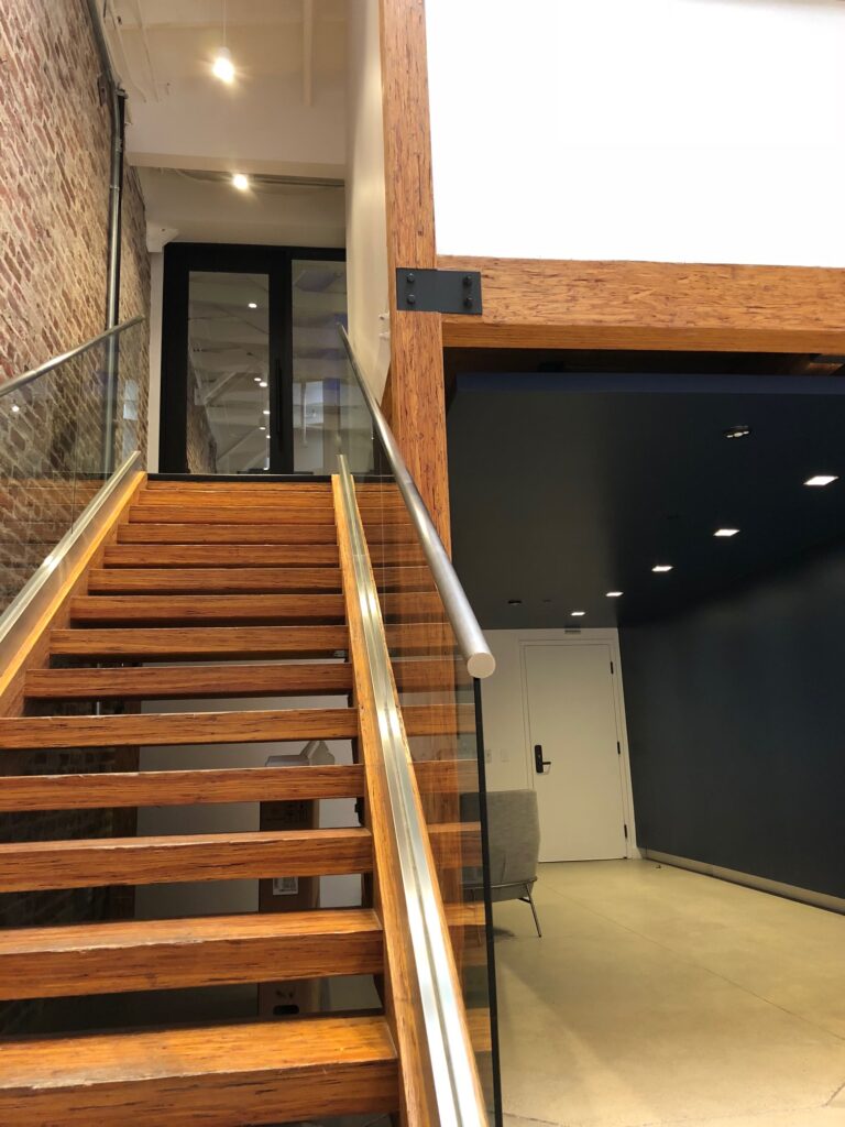 Wooden staircase with glass railing leads to a door on the upper level. The wall to the left is exposed brick. On the right, a dark hallway with a white door at the end. Ceiling lights illuminate the modern interior space.