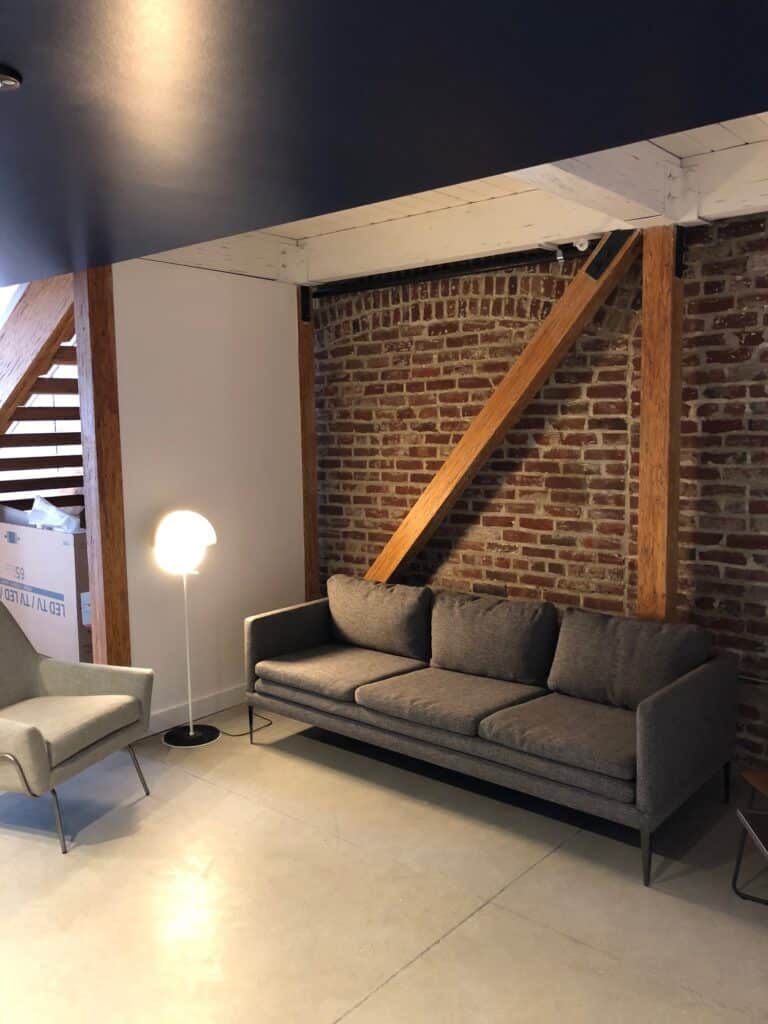 A modern living room with a gray sofa against an exposed brick wall, a small round floor lamp beside it, and a gray armchair nearby. Wooden beams and a staircase are visible in the background. The floor is light-colored concrete.