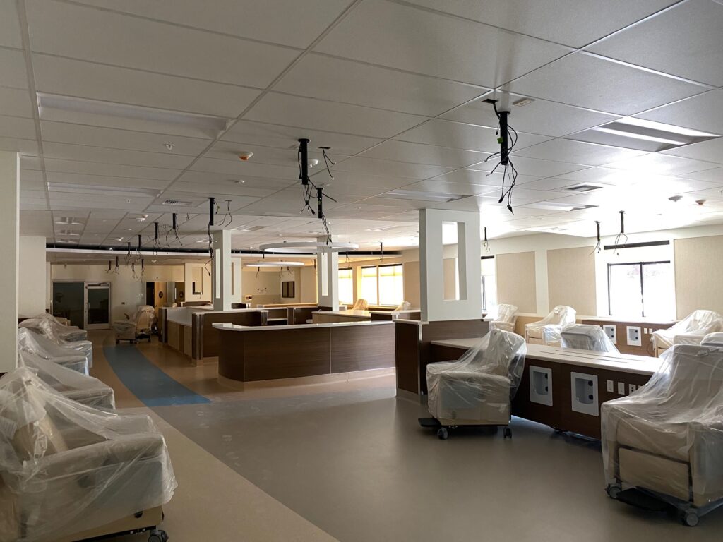A hospital room with several chairs covered in plastic, a central nurses station, and medical equipment hang from the ceiling. The room has a clean, neutral color scheme with beige walls and flooring. Natural light enters through large windows.