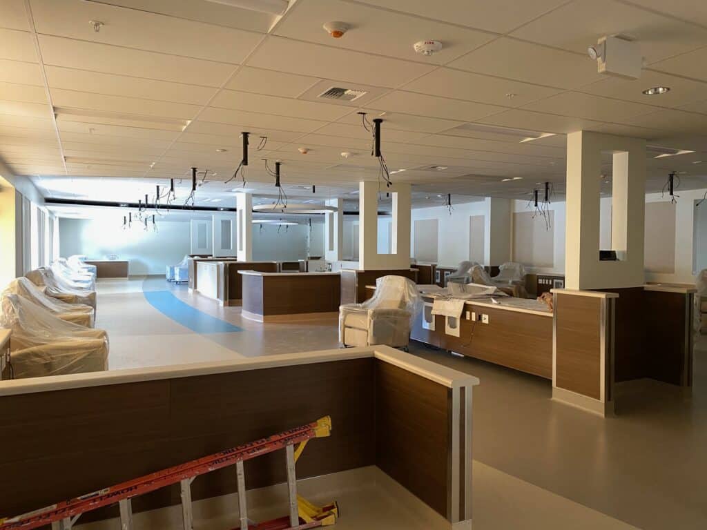 A large, empty hospital room under construction. The room features multiple cubicles with wooden partitions. Medical equipment and furniture covered in plastic. The ceiling has exposed wires and uninstalled light fixtures.