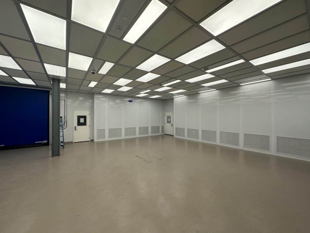 An empty, well-lit room with a beige floor and a grid-patterned ceiling featuring fluorescent lights. The walls are white with a few ventilation grilles. A blue wall or curtain is partially visible on the left side.