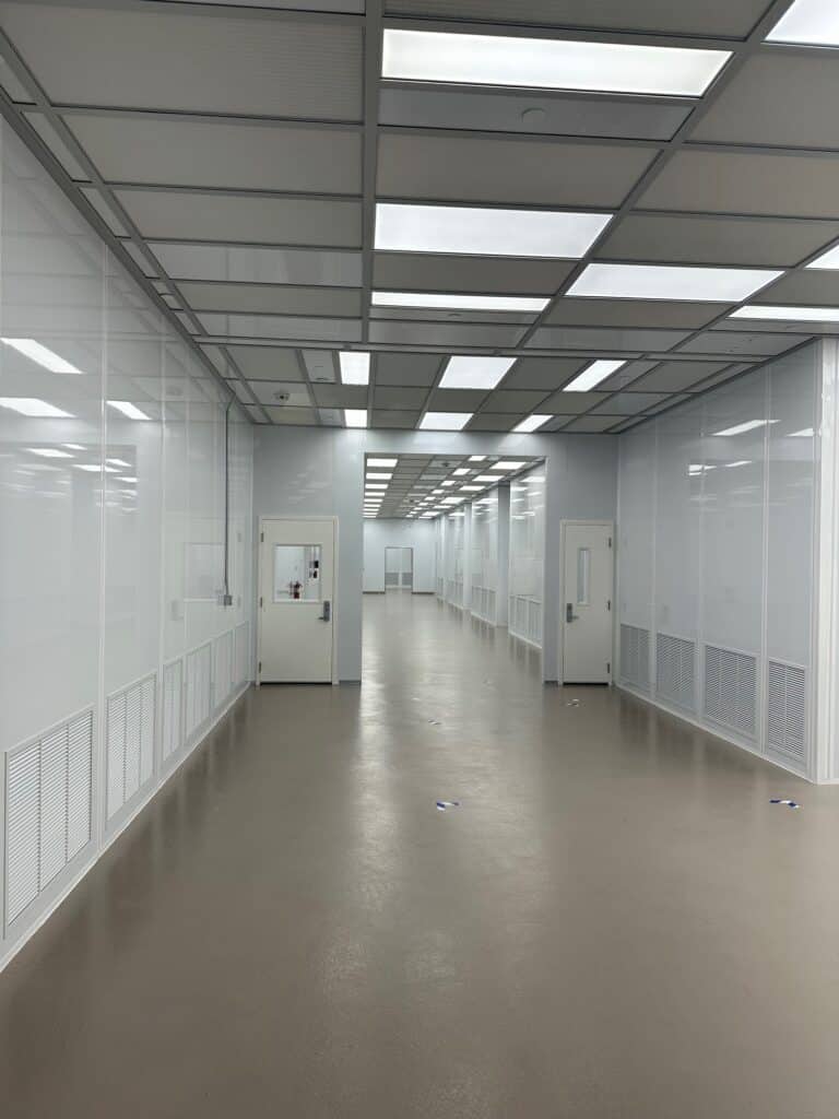 A long, empty corridor with a polished beige floor and glossy white walls. The ceiling is lined with fluorescent lights, and there are several closed white doors along the hallway. The space is bright and sterile, resembling a lab or clean room.