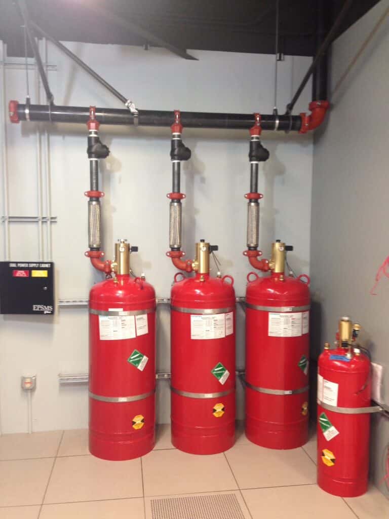 A row of four large red fire suppression cylinders with control panels and piping connections on top, next to a smaller red cylinder. The setup is in a corner of a tiled room with a black control box on the wall.