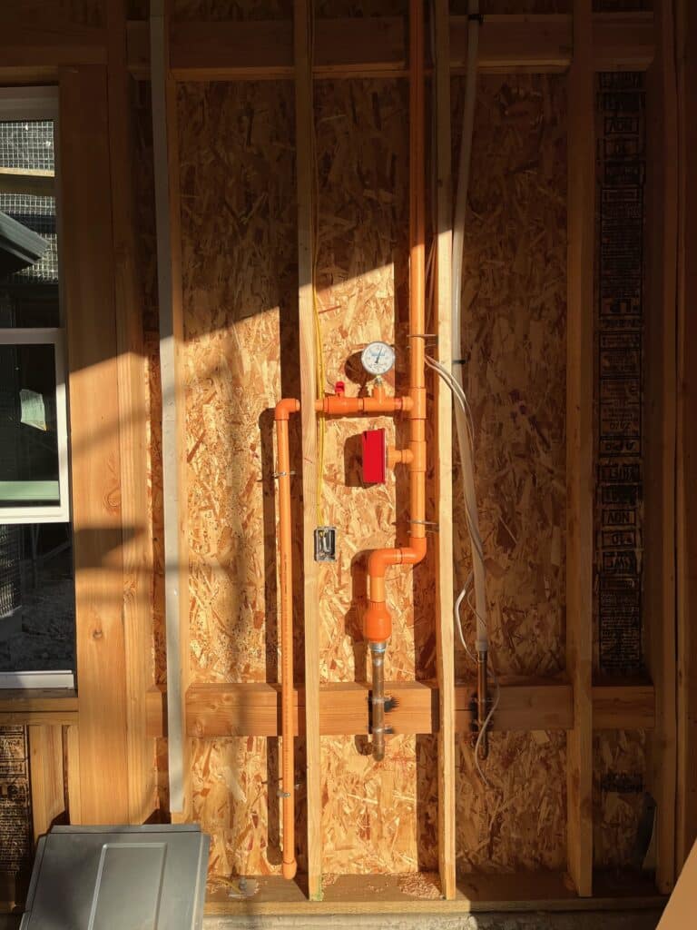 A partially constructed wall showcasing exposed orange plumbing pipes with a pressure gauge and valve, set against plywood sheathing. Sunlight casts shadows across the wooden studs, with a window partially visible on the left.
