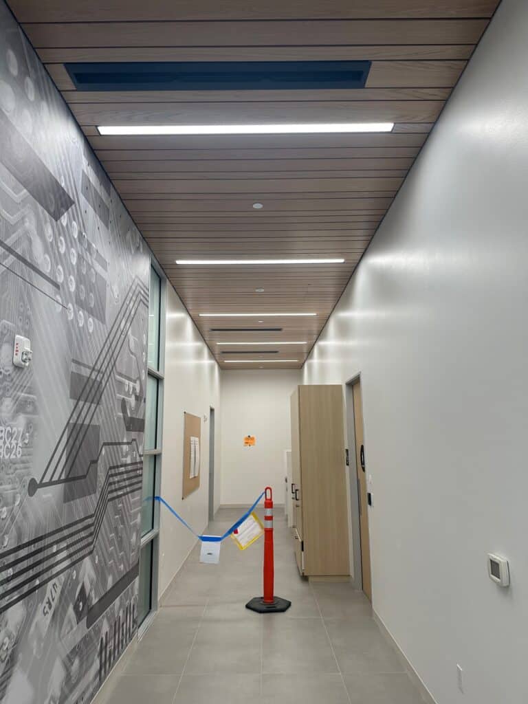 A modern hallway with a wood panel ceiling and bright lighting. A large wall graphic depicts a circuit board. The floor is tiled, and a construction cone with tape blocks access to a door. Papers are taped to a nearby closed door.