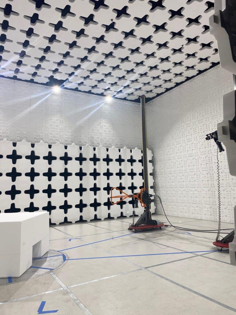 A room with white walls and ceiling, each featuring a pattern of black cross shapes. A tall pole with attached equipment stands in the corner, and a white rectangular block is in the foreground. Blue tape marks are visible on the floor.