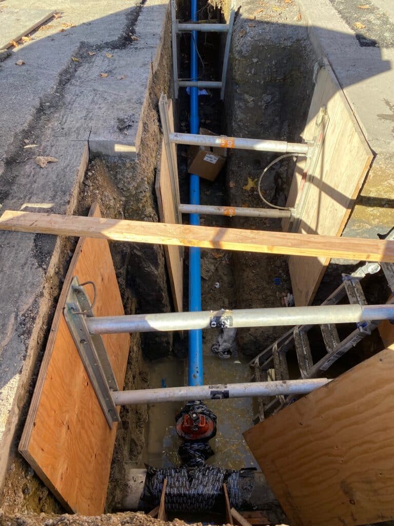 A construction trench with wooden and metal supports contains a long blue pipe. There are ladders on either side of the trench and some construction materials are visible. Sunlight casts shadows across the scene.