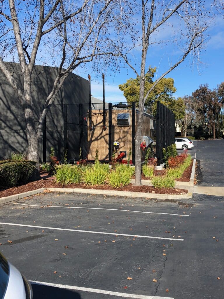 A parking lot with empty spaces next to a fenced area containing plants and two red benches. Leafless trees cast shadows on the ground. The sky is clear and blue, indicating a sunny day.
