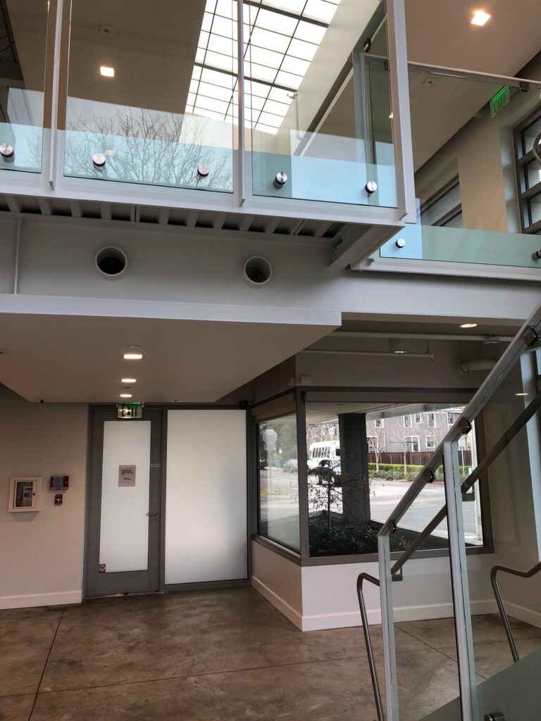 A modern, two-story interior with glass railings on an upper walkway. The ground floor has concrete flooring and a door leading outside. Large windows provide natural light, and minimalistic design elements are present throughout the space.
