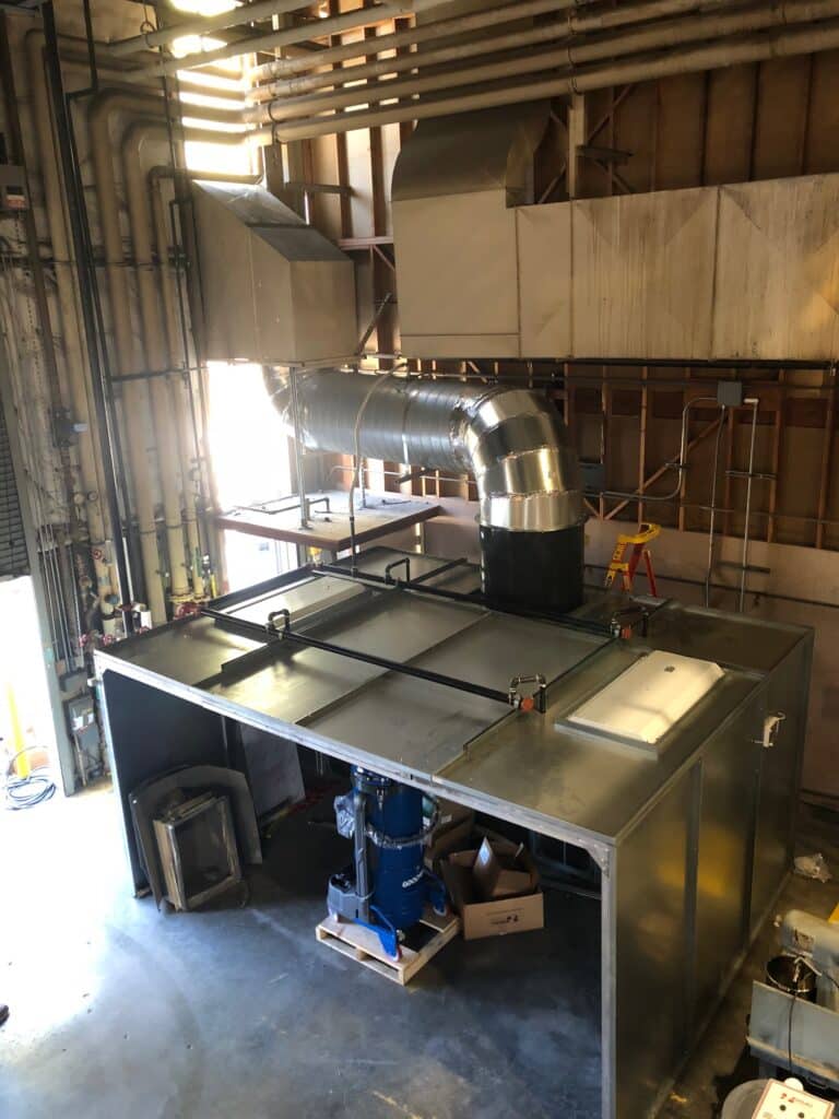 View of an industrial room featuring metal ductwork and ventilation system. The room includes a large metallic structure with pipes and equipment parts, like fans and air ducts, connected above. Various tools and boxes are on the floor.