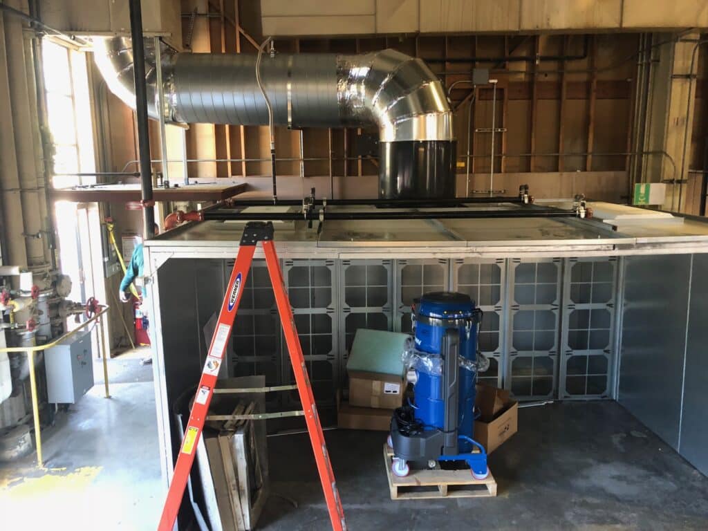 A large industrial room with a red ladder in the foreground. Behind it, theres ductwork and a ventilation system on top of a metal structure. A blue machine, possibly a compressor or pump, is on a pallet next to a cardboard box.