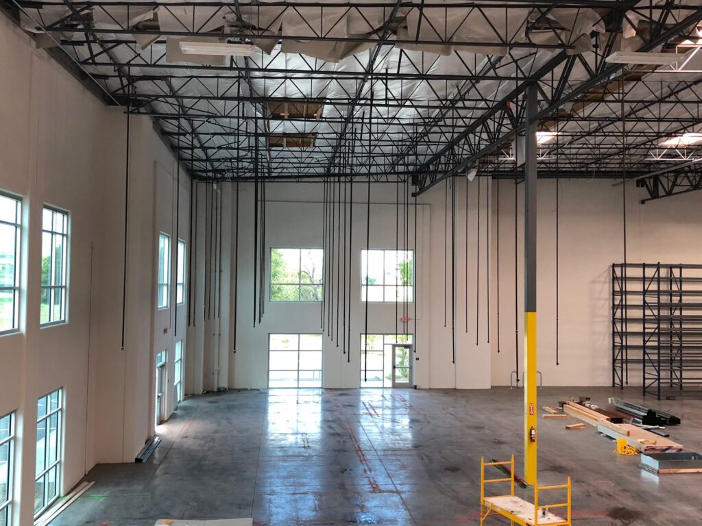 Interior of a large warehouse under construction, featuring high ceilings with visible beams and exposed wiring. Windows line the walls, allowing natural light to illuminate the concrete floor. Scaffolding and construction materials are scattered throughout.