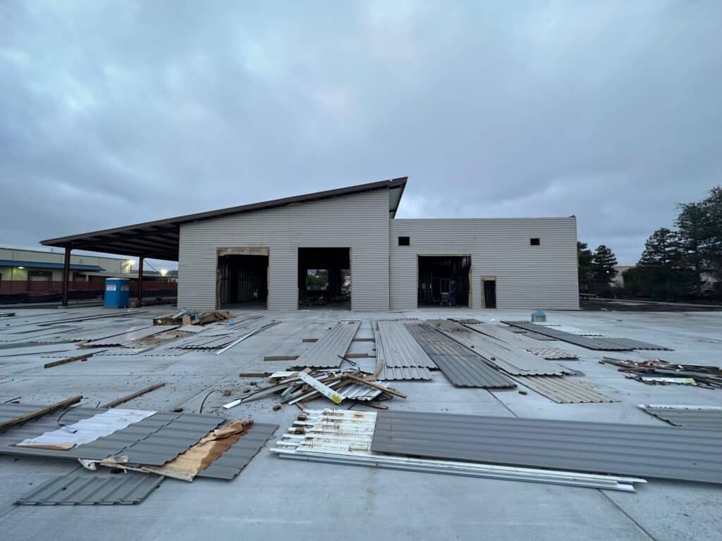 Partially constructed industrial building with an angled roof. The exterior is incomplete, with several metallic panels lying scattered on a concrete lot in front. A portable toilet is visible on the left. Overcast sky in the background.
