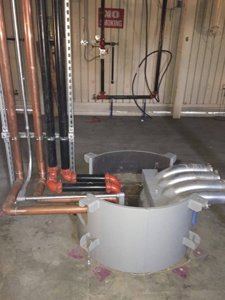 An indoor industrial setting showing metal and copper pipes arranged vertically and horizontally. They enter a large semi-circular gray enclosure on the floor. A No Smoking sign is visible on the wall behind the pipes.