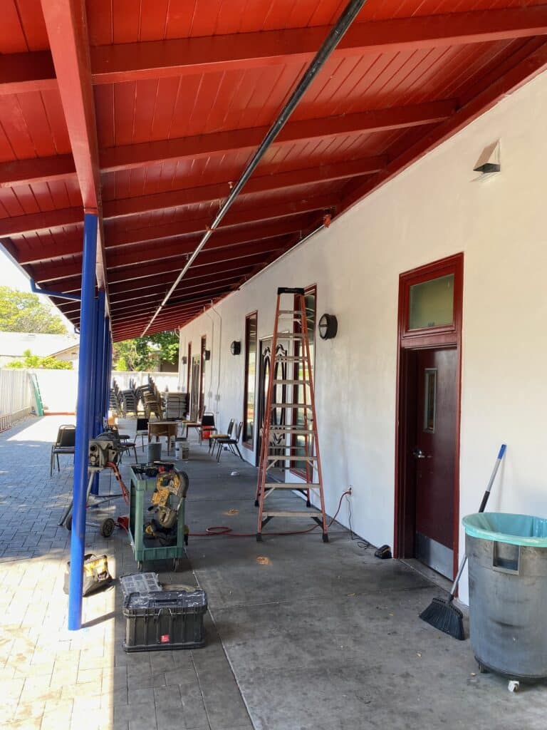 A covered outdoor walkway features a white building with a red roof. Chairs and tables are scattered along the side. A red ladder leans against the wall near a door. Various tools, a broom, and a garbage can are on the concrete ground.