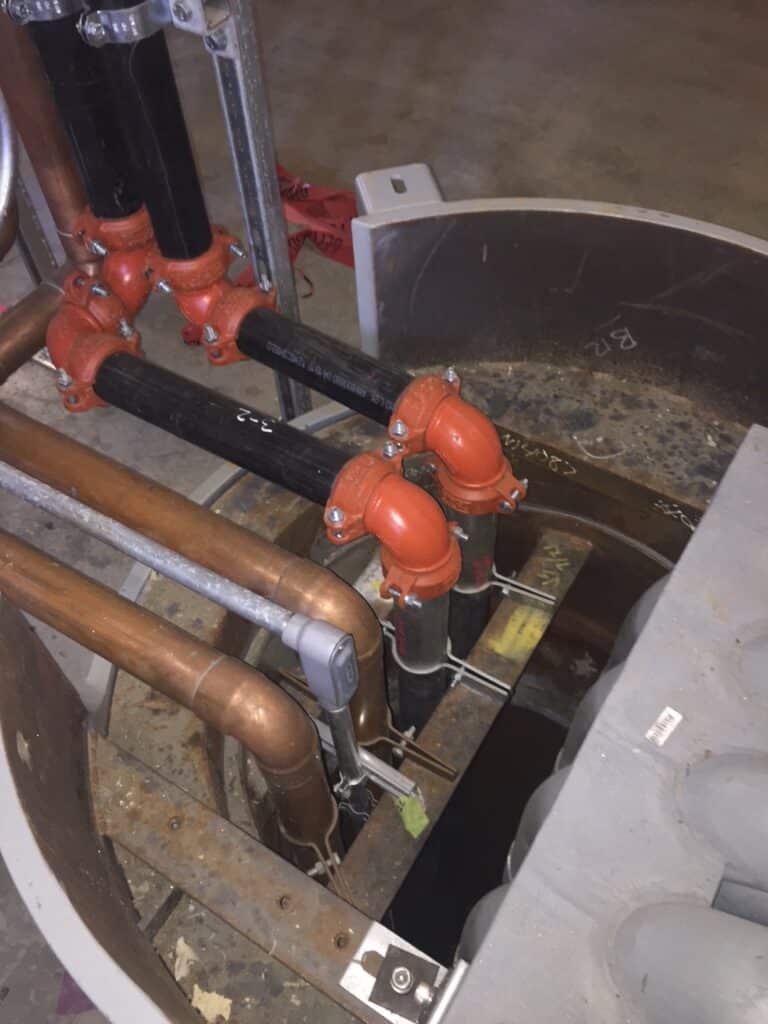 A close-up of an industrial setup with several black and copper pipes connected by orange joints, leading into a circular opening. The floor is concrete, and there are metal frames nearby. The area appears unfinished or under construction.
