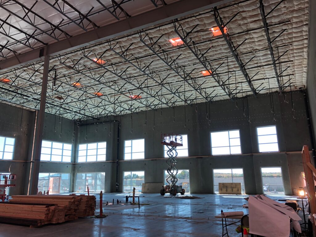 Interior of a large industrial building under construction with a high ceiling and exposed insulation. Large windows provide natural light. A scissor lift is in the center, surrounded by building materials and safety cones.