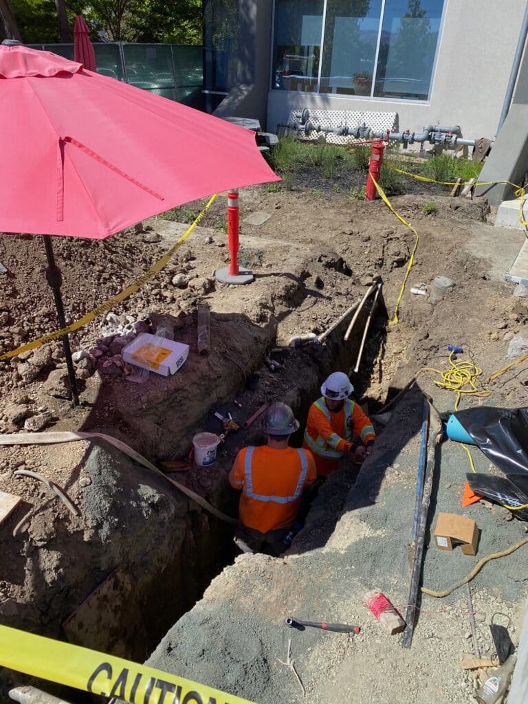 Two construction workers wearing helmets and orange vests are working in a trench. Theres caution tape around the site, a red umbrella providing shade, and construction tools and equipment are visible nearby.