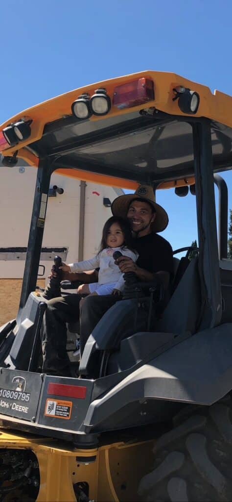 A man wearing a wide-brimmed hat is sitting in a tractor cab, smiling and holding a young child on his lap. The child is also smiling. They are outside on a sunny day, with a building and some greenery visible in the background.