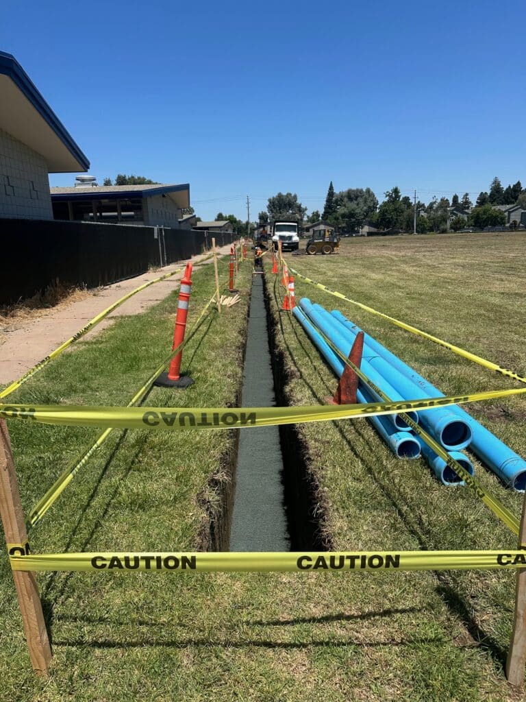 A long trench is dug into a grassy field, flanked by yellow caution tape and orange cones. Blue pipes lie beside the trench. Construction equipment is visible in the background under a clear blue sky.