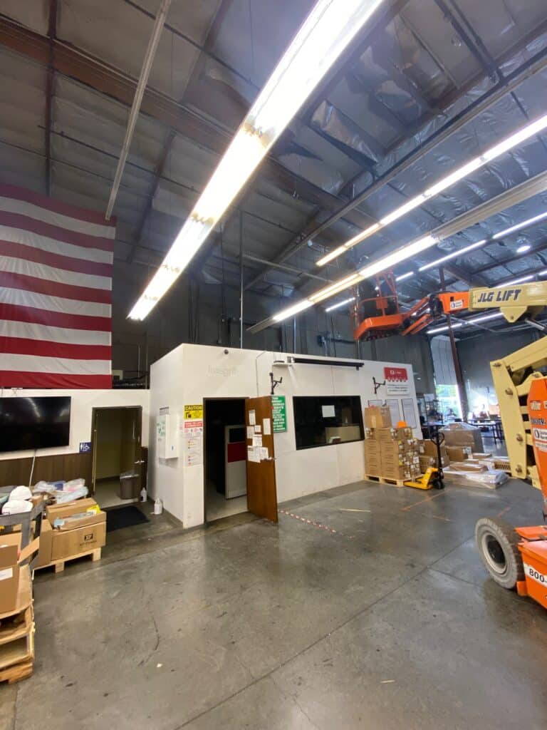 A spacious warehouse with a large American flag on the wall. There are various boxes and supplies on the floor. An orange JLG lift extends near the ceiling. Fluorescent lights illuminate the area, and a small office structure is in the center.