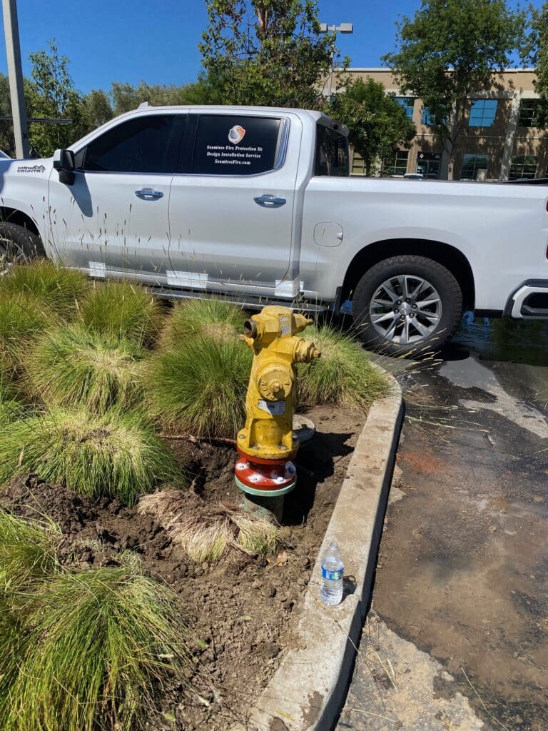 A white truck is parked closely to a yellow fire hydrant in a parking lot. Water is pooling near the curb, and a plastic water bottle is on the ground beside some green bushes. A building is visible in the background.