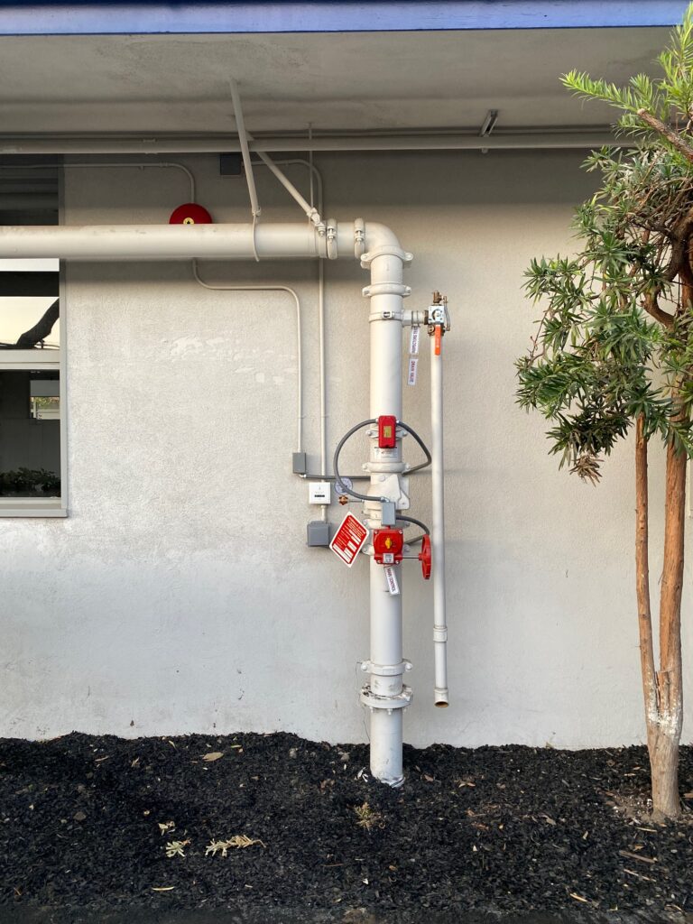 A vertical pipe system with multiple valves and red labels is mounted on an exterior wall. It is situated beside a window and near a bushy tree, with fresh mulch on the ground beneath.